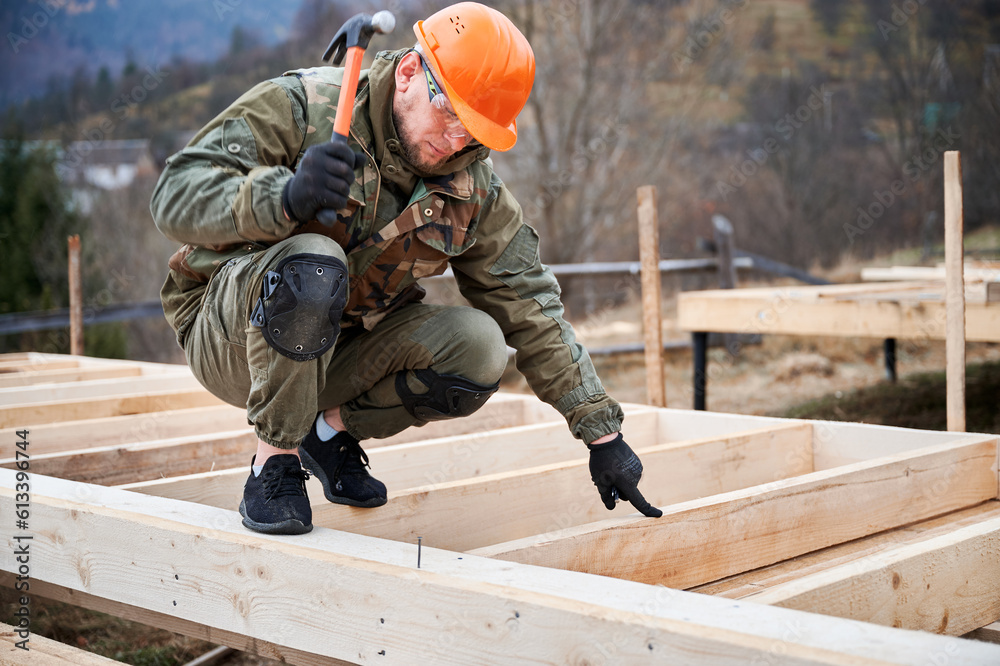 Man worker building wooden frame house on pile foundation. Carpenter hammering nail into wooden plank, using hammer. Carpentry concept. Man worker building wooden frame house on pile foundation. Carpenter hammering nail into wooden plank, using hammer. Carpentry concept.