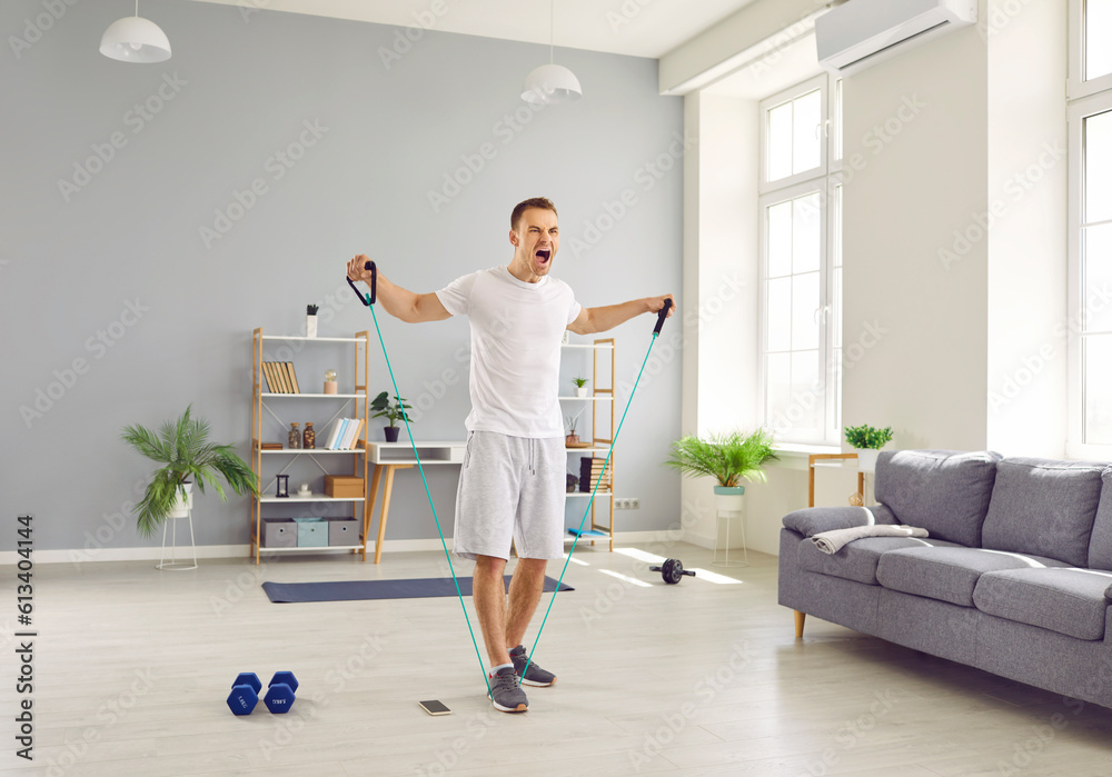 Exited young sporty man in homewear doing stretching exercises with rubber band and shouting with open mouth. Athletic guy enjoy exercising. Fitness, workout sport and home training concept. Exited young sporty man in homewear doing stretching exercises with rubber band and shouting with open mouth. Athletic guy enjoy exercising. Fitness, workout sport and home training concept.