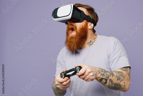 Young fun redhead bearded man he wear violet t-shirt casual clothes hold in hand play pc game with joystick console isolated on plain pastel light purple background studio portrait. Lifestyle concept.