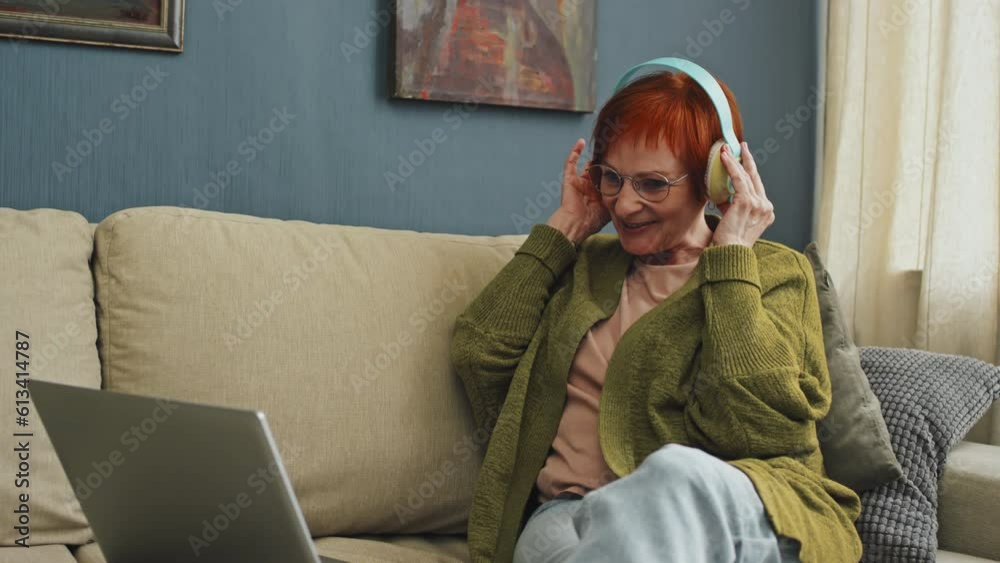 Tilt shot of smiling senior woman wearing headphones and glasses sitting on sofa with laptop waving hand and talking to family during video call Tilt shot of smiling senior woman wearing headphones and glasses sitting on sofa with laptop waving hand and talking to family during video call
