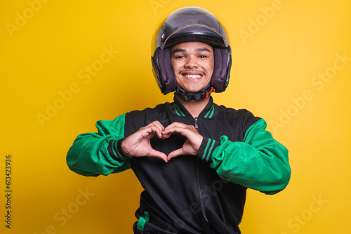Asian online taxi driver wearing green jacket and helmet showing heart sign over yellow background