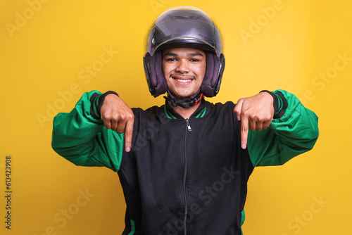 Asian online taxi driver wearing green jacket and helmet pointing fingers down over yellow background