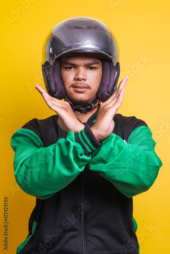Asian online taxi driver wearing green jacket and helmet crossing hands, gesturing say no warning or prohibition against yellow background