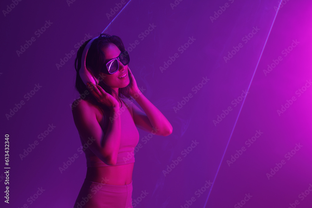 Fototapeta premium Beautiful woman with purple hair in futuristic costume over dark background. Violet neon light. Portrait of young girl in modern headphones listening music.