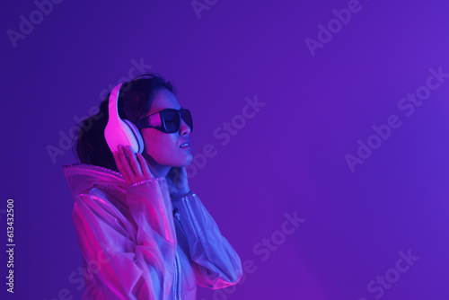 Beautiful woman with purple hair in futuristic costume over dark background. Violet neon light. Portrait of young girl in modern headphones listening music.