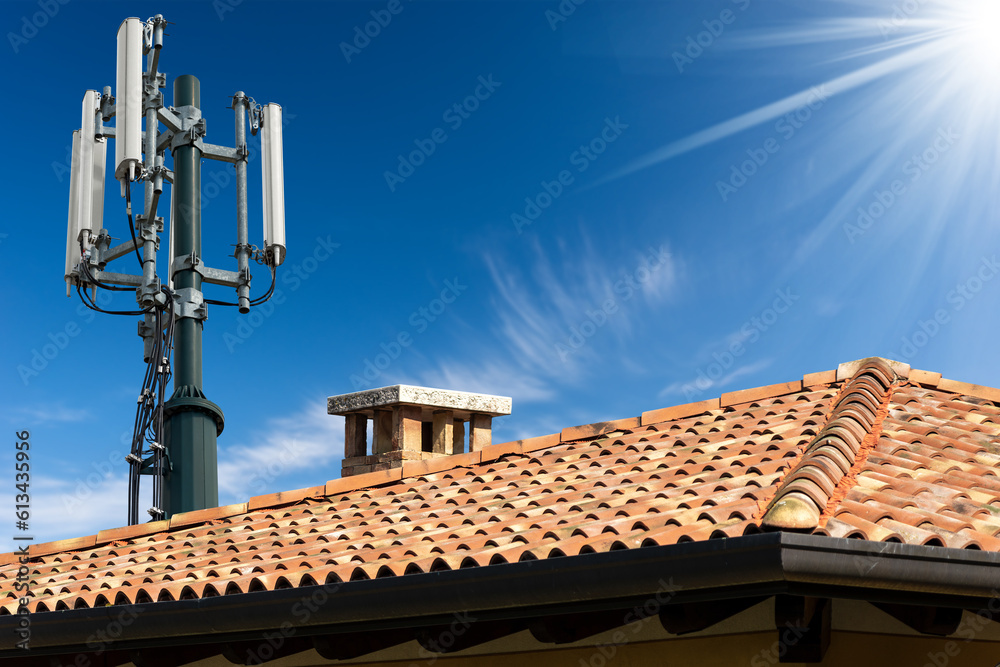 Closeup of a house roof with a telecommunications tower with antennas (aerial), against a clear blue sky with clouds and copy space.