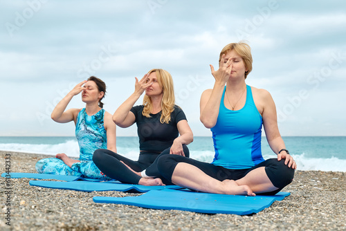 Summer pilates training. Group of women practicing yoga exercise at the beach, doing breath practice lotus pose near the water. Yoga retreat workout, body and mind wellness at the sea.