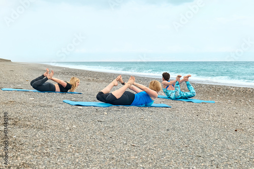 Summer pilates training. Group of women practicing yoga exercise at the beach near the water. Yoga retreat workout, body and mind wellness at the sea.