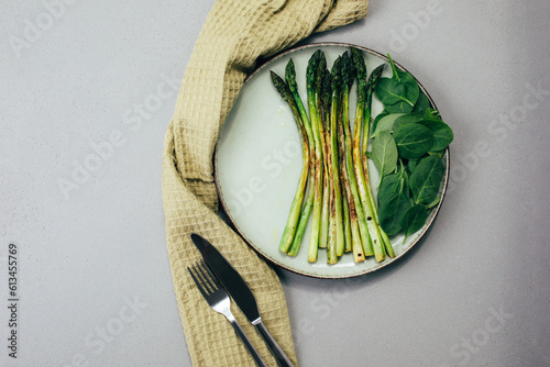 Cooked green asparagus on a green plate with fresh spinach leaves on a gray table with a green kitchen towel. Healthly food. Top view