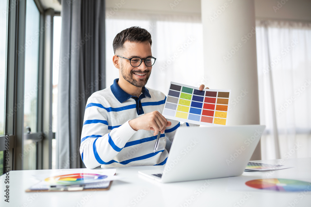 Cheerful man designer having video conference with clients, sitting at desk in front of computer, holding color palettes, gesturing and smiling, copy space Cheerful man designer having video conference with clients, sitting at desk in front of computer, holding color palettes, gesturing and smiling, copy space
