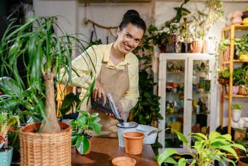 Happy smiling female shop worker pouring soil, getting ready to transplant plants in a cozy plant shop. Seedlings planting process. Home gardening. Hobbies and leisure concept.