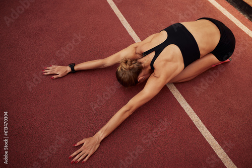 A young athlete girl is warming up before the race at the stadium. A slender blonde woman in a black tracksuit kneads her muscles sitting on her knees. Sports and recreation. Top view.