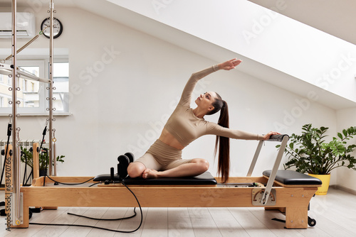 A young girl is doing Pilates on a reformer bed in a bright studio. A slender brunette in a beige bodysuit makes bends to strengthen the spine and back muscles. Healthy lifestyle concept.