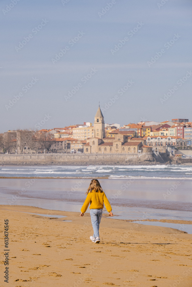 child girl walking on the beach with the town of Gijon, Asturias, in the background. She is wearing winter clothes, jumper and jeans. child girl walking on the beach with the town of Gijon, Asturias, in the background. She is wearing winter clothes, jumper and jeans.
