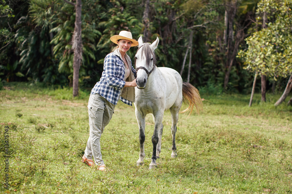 Young woman in shirt and straw hat posing next to white gray horse, jungle trees background - horseriding, ranch at Isalo Park, Madagascar