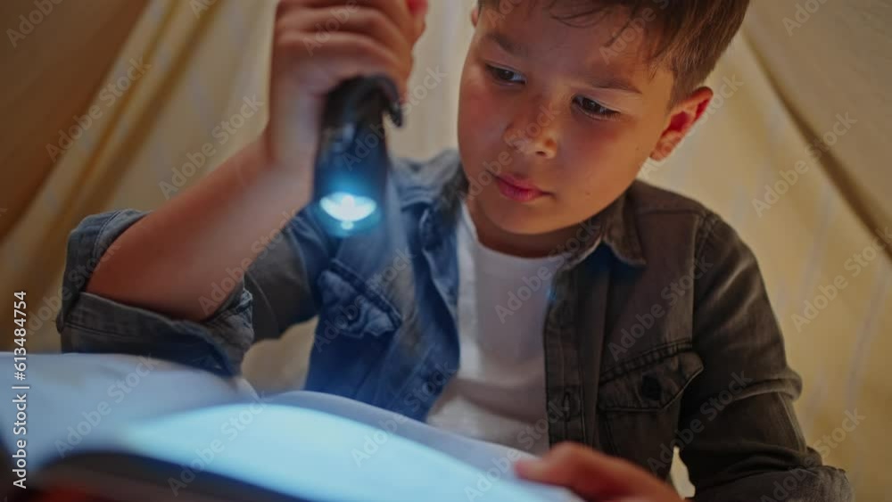 Close-up emotional school boy reading fairy tales with flashlight, hiding inside a lit tent wigwam Close-up emotional school boy reading fairy tales with flashlight, hiding inside a lit tent wigwam