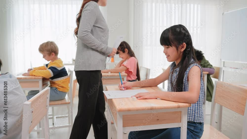 Group diverse students learning with woman teacher at elementary School.Young teacher woman handing out papers for exams to school children sit on desk in classroom at international school Group diverse students learning with woman teacher at elementary School.Young teacher woman handing out papers for exams to school children sit on desk in classroom at international school