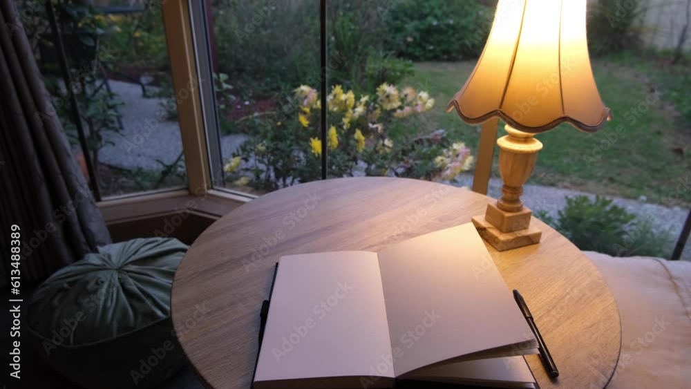 An empty open notebook and retro style lamp on a table by the window with a garden view. Blank mockup template of a book in room with a warm and cozy atmosphere at home.