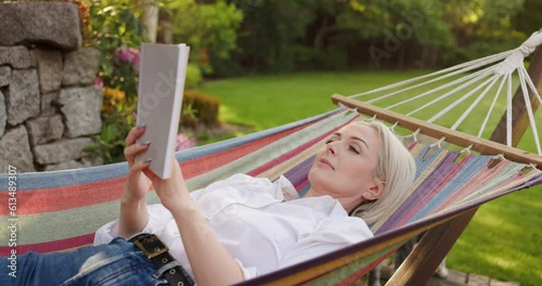 Adult woman reading book while lying in hammock in backyard garden