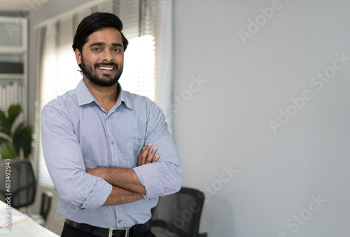 Portrait of indian businessman happy standing arms crossed in modern office. Confident young smart bearded man entrepreneur looking at camera smiling. Friendly india ethnic manager work in workplace.