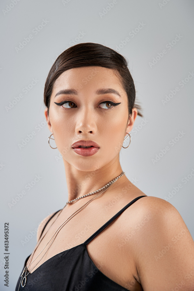 portrait of alluring asian woman in elegant black dress, silver necklaces and earrings, with bold makeup and brunette hair looking away on grey background, fashionable spring portrait of alluring asian woman in elegant black dress, silver necklaces and earrings, with bold makeup and brunette hair looking away on grey background, fashionable spring