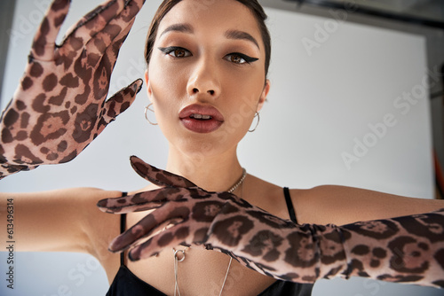portrait of elegant asian woman with bold makeup and expressive gaze, in black strap dress and animal print gloves looking at camera on grey background, spring fashion photography
