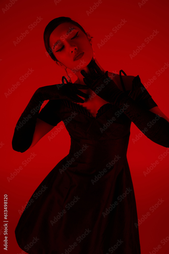 Fototapeta premium expressive and graceful asian woman in black long gloves and elegant cocktail dress touching neck and posing on vibrant background with red lighting effect, spring fashion photography