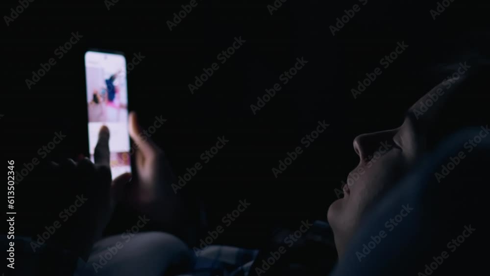 The girl lies in bed and looks into the bright screen of the smartphone addiction to social networks. Smartphone in hand. Woman looking at phone screen at night while studying social media news
