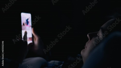 The girl lies in bed and looks into the bright screen of the smartphone addiction to social networks. Smartphone in hand. Woman looking at phone screen at night while studying social media news
