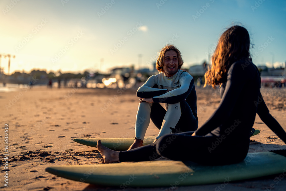 Two surfers with surfboard sitting on beach resting after surfing at sunset.