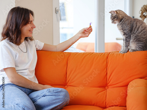 Canvas Print Young woman is playing with her gray cat on orange sofa at home