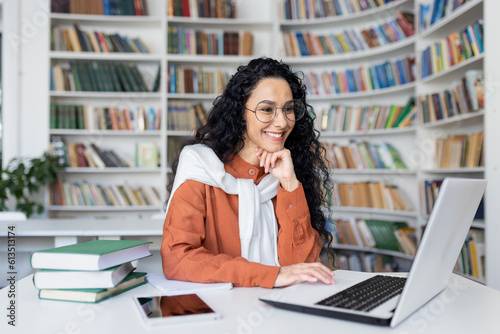 Female student with laptop reading online video course, Latin American woman smiling and satisfied with independent online learning sitting inside university campus in library. © Liubomir