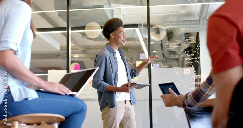 Happy biracial casual businessman presenting to diverse colleagues at glass wall, slow motion