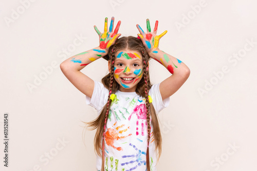 A little schoolgirl with painted hands and face with multicolored paints. A girl stained with paints. The concept of children's creativity. Funny child shows horns. White isolated background.