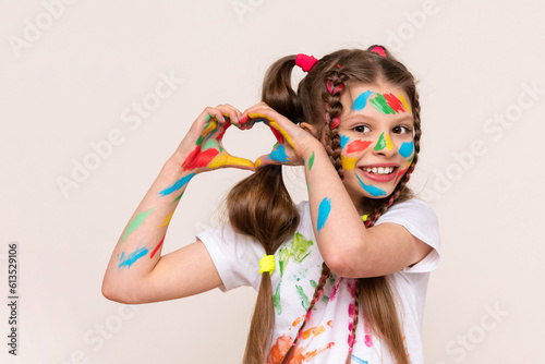 A little schoolgirl with painted hands and face with multicolored paints. A girl stained with paints. The concept of children's favorite creativity. Funny baby shows heart. White isolated background.