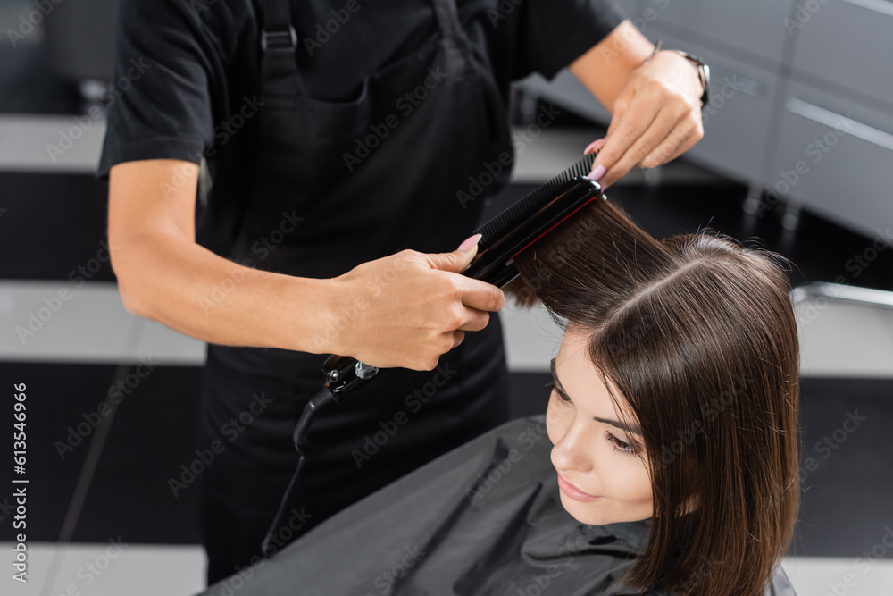 Fototapeta premium top view of professional hairdresser with hair straightener styling hair of female customer, beauty worker, client satisfaction, brunette woman with short hair, beauty salon, hair fashion