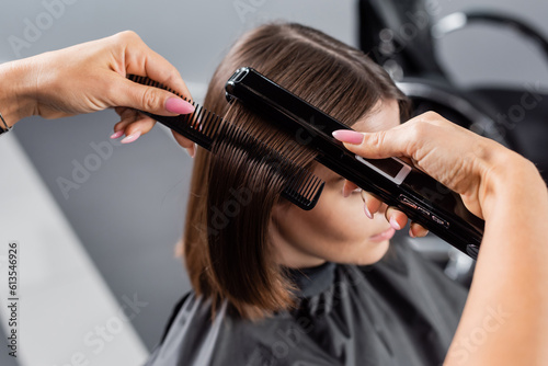 beauty profession, straightening hair, hairdresser with braids brushing and styling short brunette hair of woman in cape, hair smoothing, professional, beauty salon work, hair salon, high angle