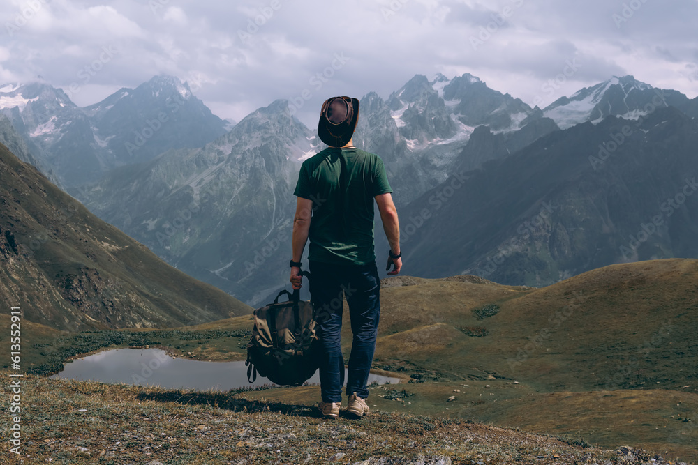 Fototapeta premium A man traveler with backpack and leather hat near the Koruldi Lake, Mestia in the Greater Caucasus Mountain Range Upper Svaneti, Georgia. Wildlife, travel and vacation. Summer day. Rear view