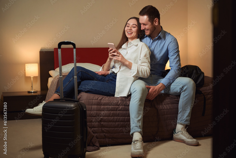 Couple browsing phone and laugh sitting on bed in hotel Couple browsing phone and laugh sitting on bed in hotel