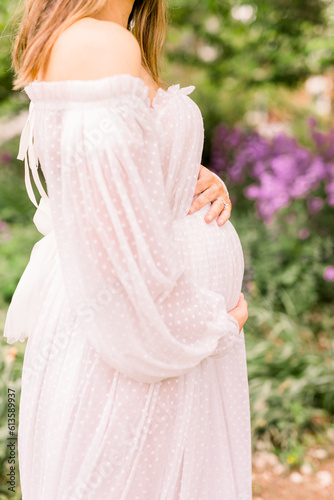 A side view of an expectant mother cradling her belly as she stands in front of greenery and purple flowers.
