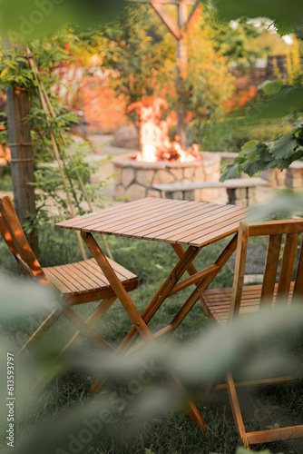 Wooden furniture in the garden against the backdrop of a fire on the patio. Among the branches of the bush stands a wooden table and chairs.