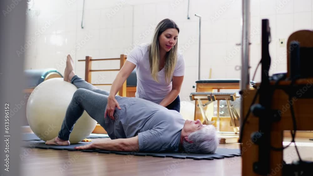 Physiotherapist guiding senior woman to exercise with Pilates ball. An elderly lady laid on floor in workout routine with the help of a sport coach
