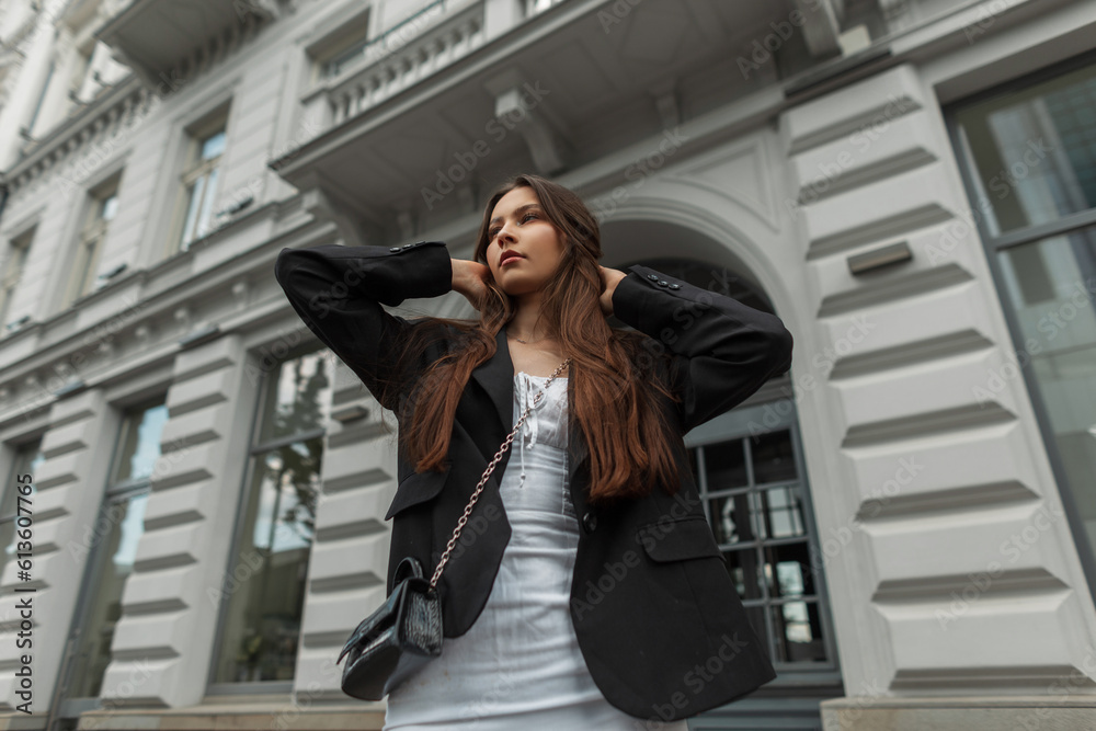 Fototapeta premium Beautiful young stylish girl model in a white dress with a black blazer with a handbag walks in the city near a vintage building