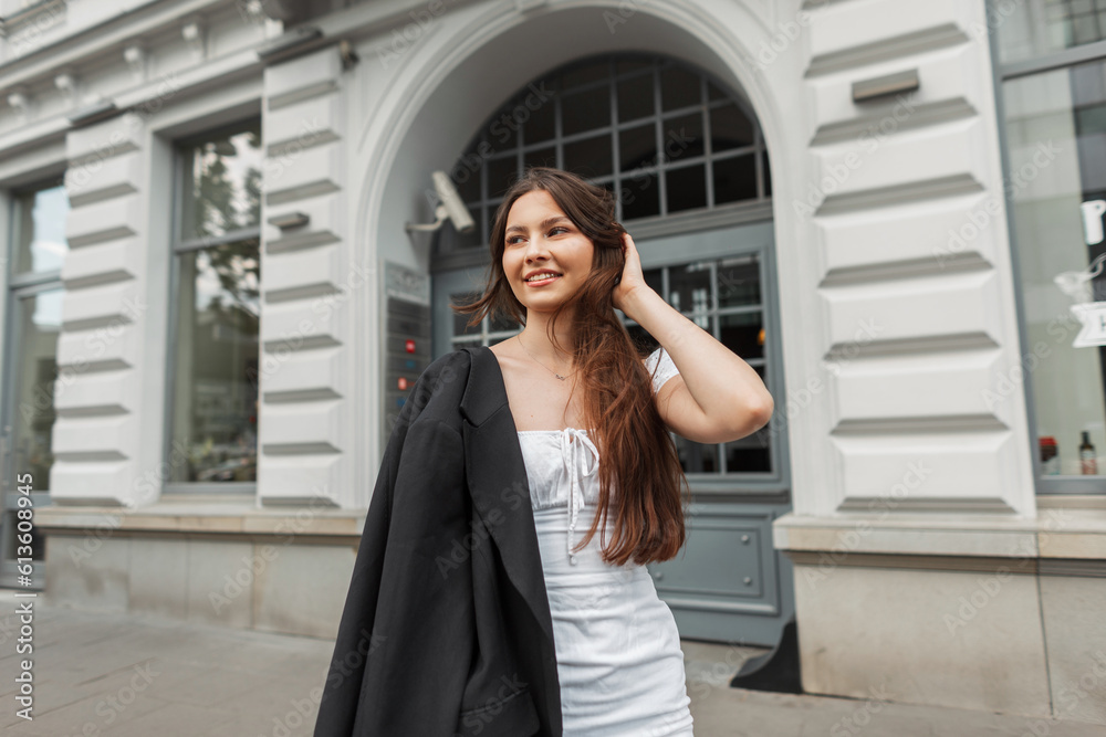 Fashionable beautiful young happy girl with a smile in stylish clothes with a white dress and a black blazer walks in the city Fashionable beautiful young happy girl with a smile in stylish clothes with a white dress and a black blazer walks in the city