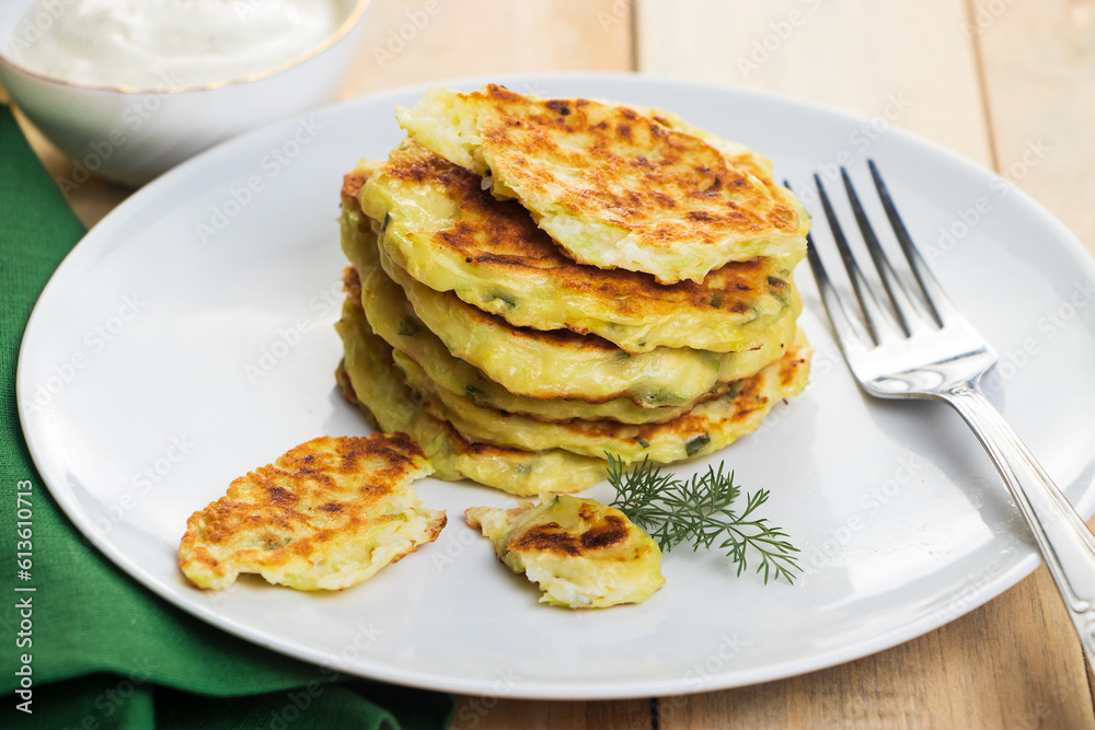 Stack of zucchini fritters in plate and cream sauce. Dish on green napkin. Healthy vegan diet food on wooden background.