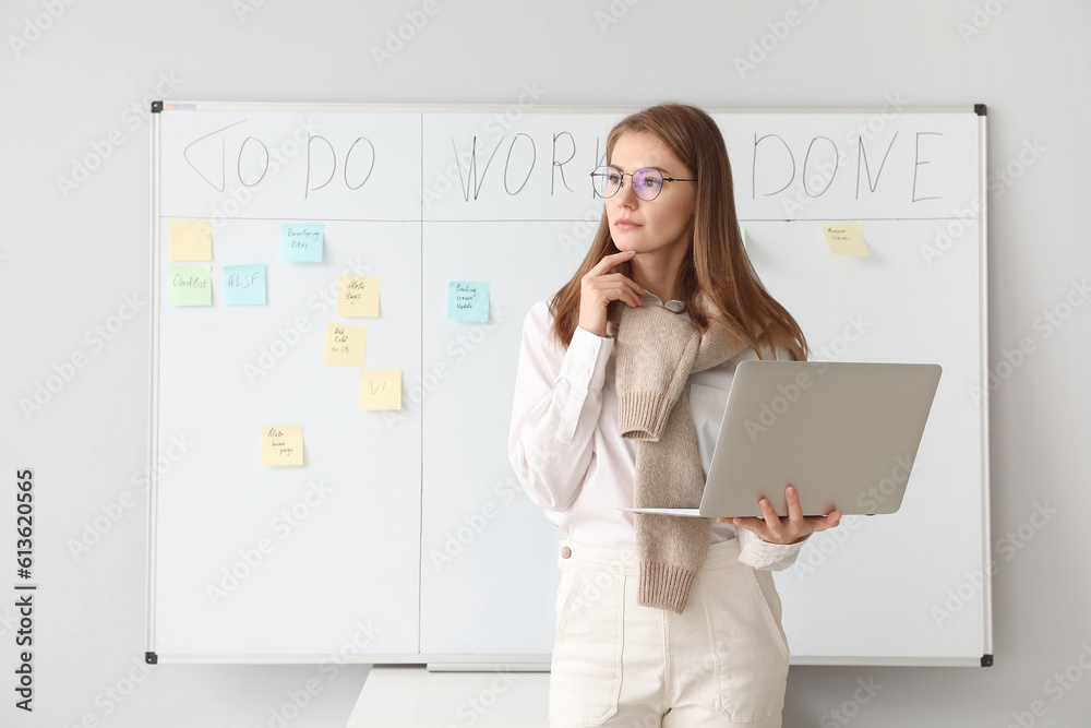 Thoughtful young businesswoman with laptop near scrum task board in office Thoughtful young businesswoman with laptop near scrum task board in office