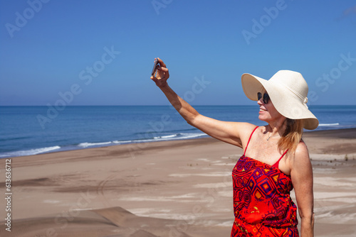 Happy mature woman smiling while taking selfies with mobile phone on top on beach dune. Summer and technology concept.