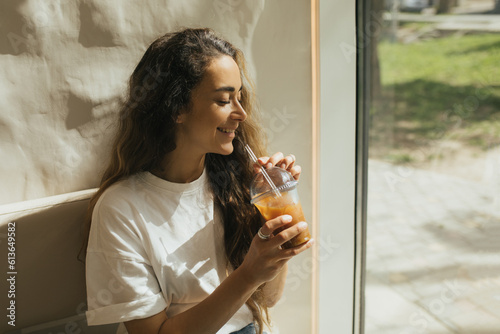 Young woman with long curly hair drinking iced coffee on beige background.