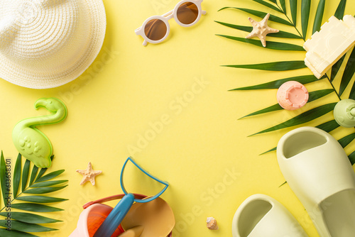 Tropical resort with kids. Top view photo of empty place surrounded by palm leaves and shells, sand toys, slippers with panama and sunglasses on yellow isolated background with copyspace