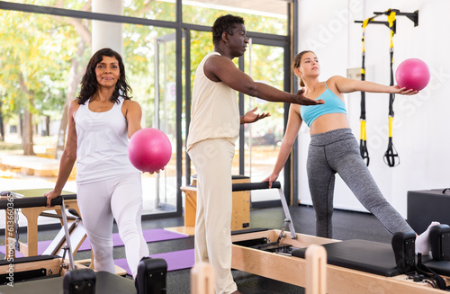 Hispanic and caucasian women exercising with pilates reformers and small fitness balls. African-american man pilates instructor assisting them with workout.